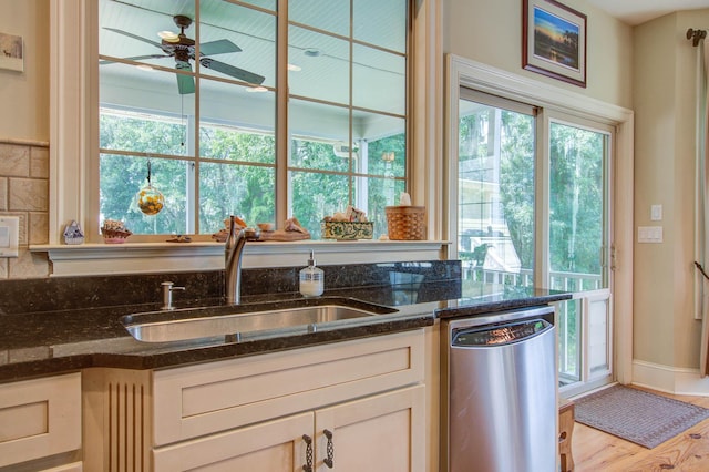 kitchen featuring stainless steel dishwasher, a sink, a ceiling fan, dark stone counters, and light wood-style flooring