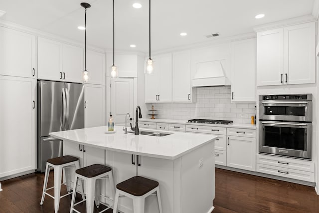 kitchen with appliances with stainless steel finishes, custom range hood, a kitchen island with sink, sink, and white cabinetry