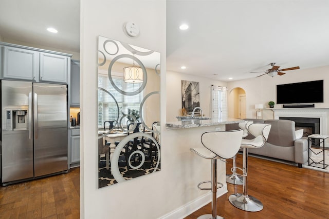 kitchen with a breakfast bar area, gray cabinetry, stainless steel refrigerator with ice dispenser, light stone countertops, and dark wood-type flooring