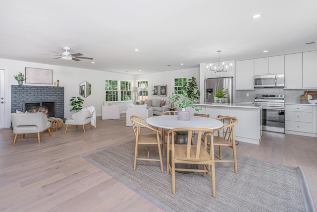 dining room featuring light hardwood / wood-style floors, ceiling fan with notable chandelier, and a brick fireplace