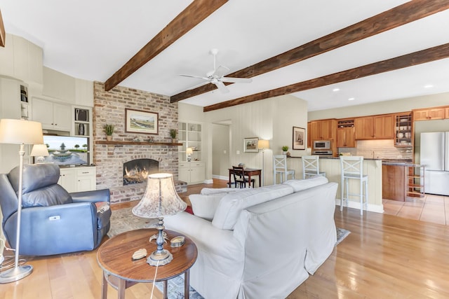 living room with a brick fireplace, beamed ceiling, light wood finished floors, and ceiling fan