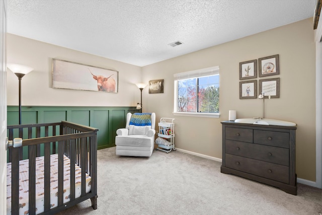 bedroom featuring carpet, a wainscoted wall, visible vents, a nursery area, and a textured ceiling