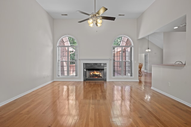 unfurnished living room featuring a towering ceiling, plenty of natural light, light wood-style floors, a ceiling fan, and a premium fireplace