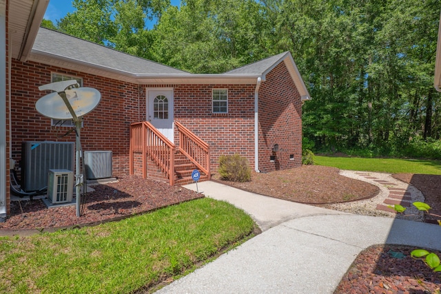 view of exterior entry with brick siding, roof with shingles, central air condition unit, and a lawn