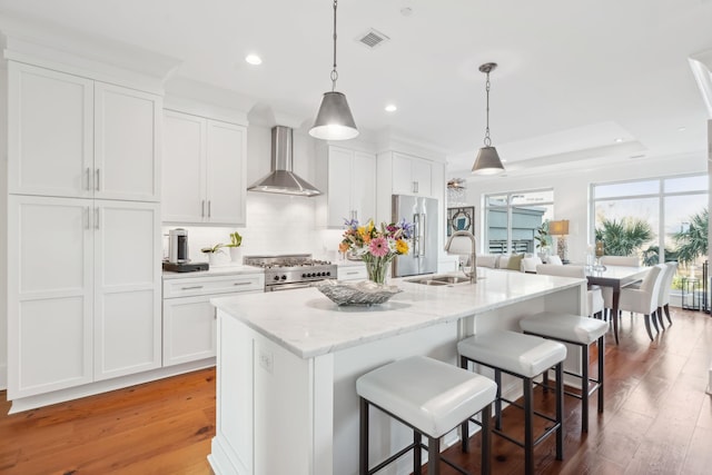 kitchen with high quality appliances, a kitchen island with sink, wall chimney range hood, white cabinetry, and a sink