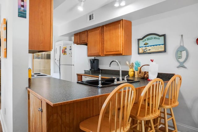 kitchen featuring a peninsula, a sink, brown cabinetry, dark countertops, and a kitchen bar