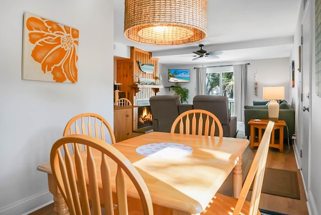 dining room featuring a ceiling fan, a lit fireplace, baseboards, and wood finished floors