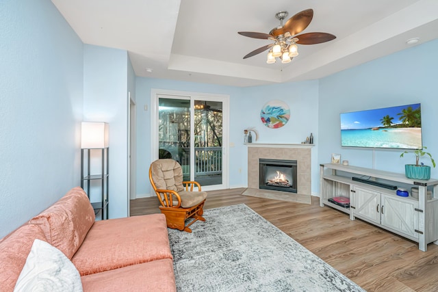 living room with a tiled fireplace, ceiling fan, light hardwood / wood-style floors, and a tray ceiling