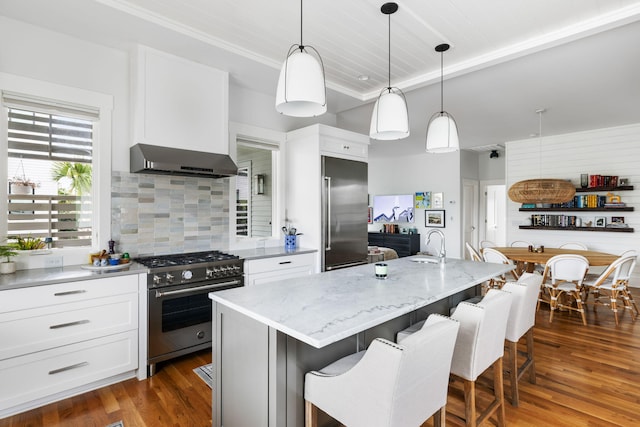 kitchen featuring pendant lighting, premium appliances, an island with sink, white cabinets, and wall chimney exhaust hood