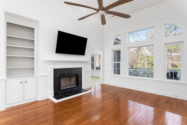 unfurnished living room featuring built in shelves, crown molding, a ceiling fan, a tile fireplace, and hardwood / wood-style floors