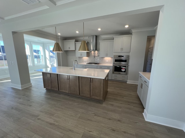 kitchen featuring a center island with sink, appliances with stainless steel finishes, decorative backsplash, decorative light fixtures, and wall chimney exhaust hood