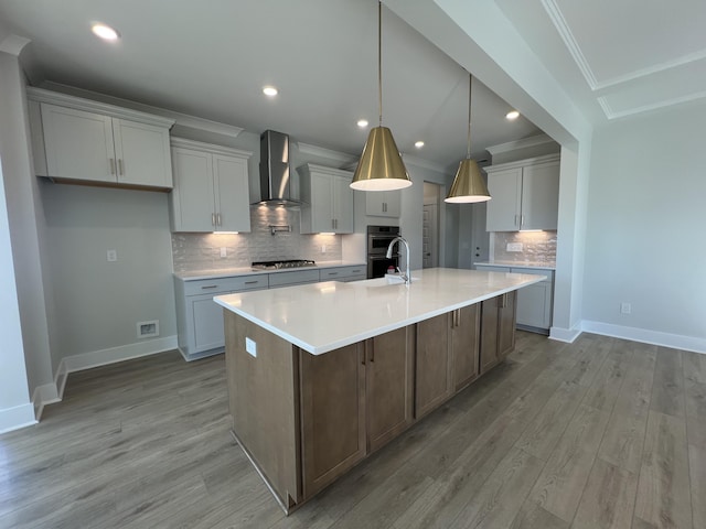 kitchen with wall chimney range hood, stainless steel appliances, an island with sink, hanging light fixtures, and light wood-type flooring