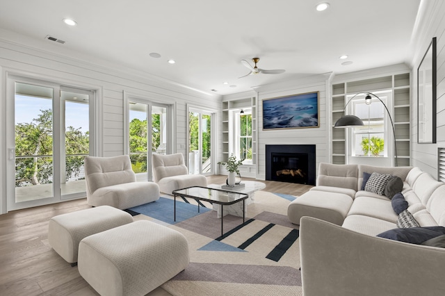 living room featuring crown molding, ceiling fan, a large fireplace, built in shelves, and light wood-type flooring