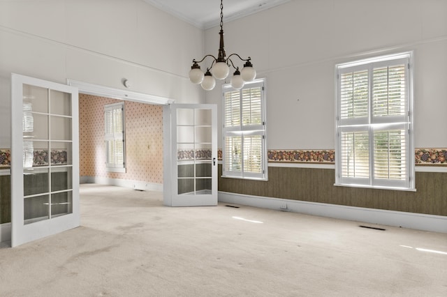 carpeted empty room featuring wallpapered walls, a chandelier, crown molding, french doors, and healthy amount of natural light