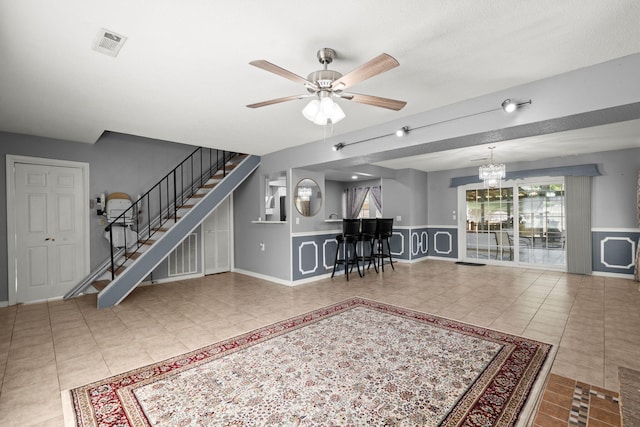 tiled living area featuring stairs, visible vents, ceiling fan with notable chandelier, and a wainscoted wall