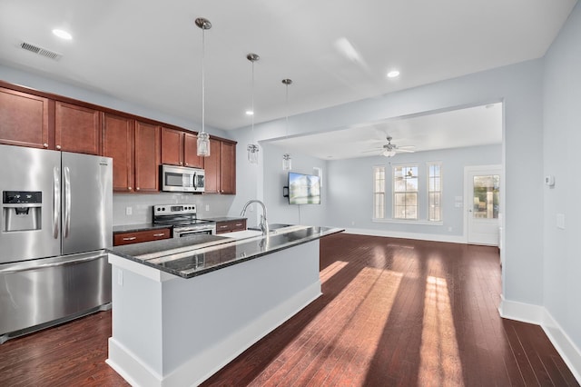 kitchen featuring appliances with stainless steel finishes, dark hardwood / wood-style flooring, dark stone counters, pendant lighting, and an island with sink