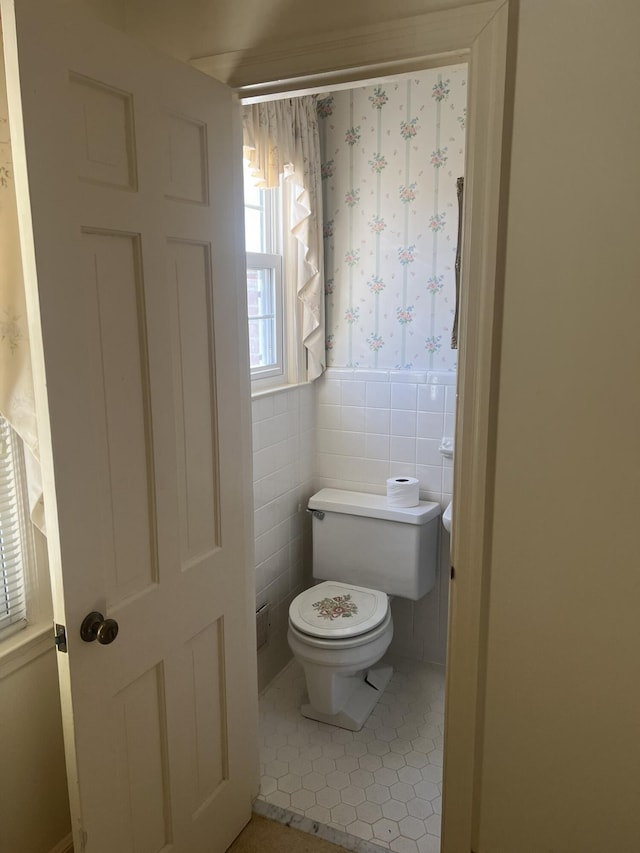 bathroom with wallpapered walls, wainscoting, toilet, tile patterned flooring, and tile walls