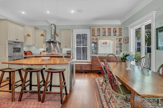 kitchen featuring wall chimney range hood, an island with sink, light wood-type flooring, and crown molding