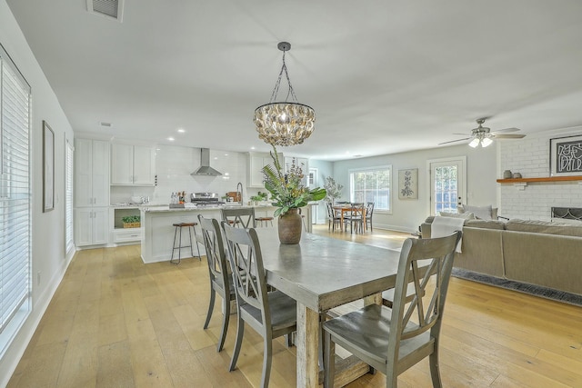 dining space with light wood-type flooring, a brick fireplace, and ceiling fan with notable chandelier