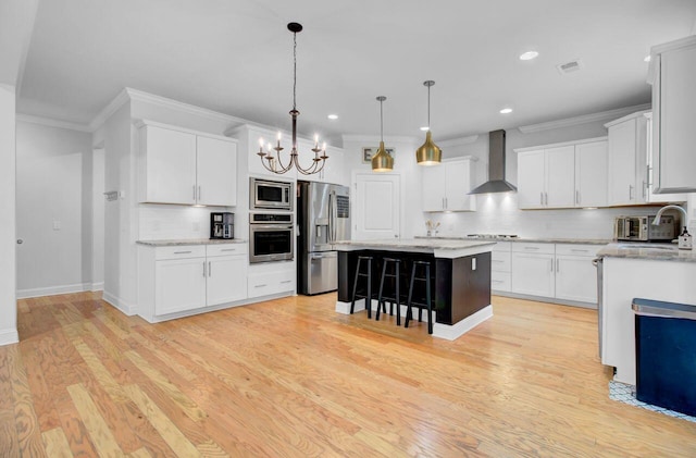 kitchen with hanging light fixtures, wall chimney exhaust hood, light wood-type flooring, appliances with stainless steel finishes, and a kitchen island