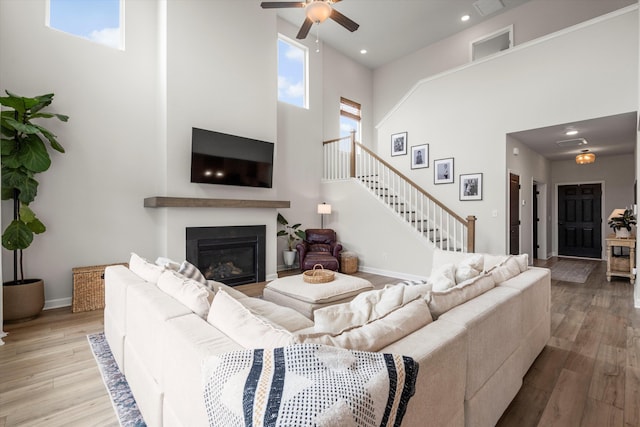 living room featuring stairs, a glass covered fireplace, wood finished floors, ceiling fan, and a high ceiling