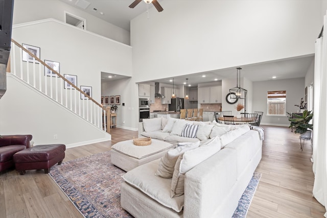 living room featuring a towering ceiling, baseboards, stairway, light wood finished floors, and ceiling fan with notable chandelier
