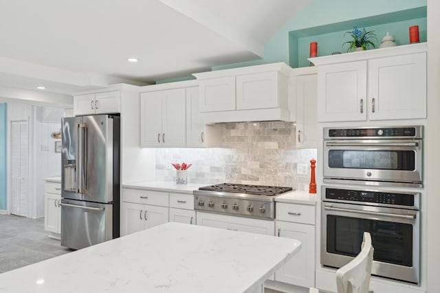 kitchen with lofted ceiling, light stone counters, stainless steel appliances, white cabinetry, and backsplash