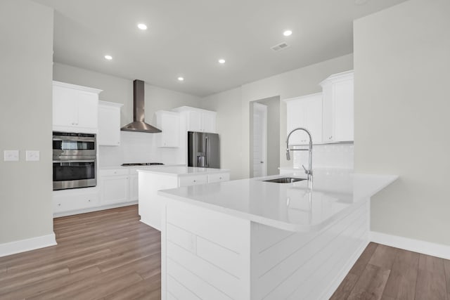 kitchen featuring light wood-type flooring, wall chimney exhaust hood, stainless steel appliances, a center island with sink, and white cabinetry