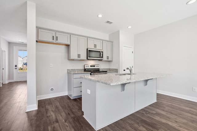 kitchen with a center island with sink, appliances with stainless steel finishes, light stone countertops, gray cabinetry, and a sink