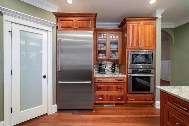 kitchen featuring light stone counters, dark hardwood / wood-style floors, built in appliances, crown molding, and decorative backsplash