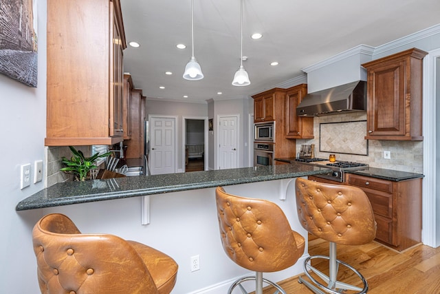 kitchen featuring a kitchen bar, decorative light fixtures, dark stone countertops, stainless steel appliances, and wall chimney range hood