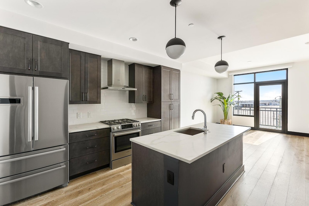 kitchen with hanging light fixtures, appliances with stainless steel finishes, sink, wall chimney range hood, and an island with sink