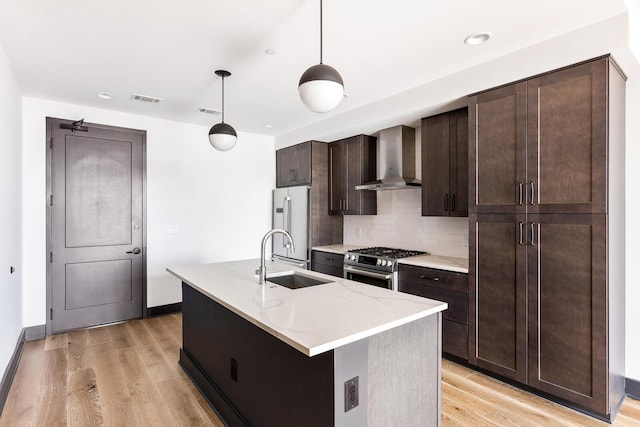 kitchen featuring appliances with stainless steel finishes, a kitchen island with sink, sink, decorative light fixtures, and wall chimney exhaust hood