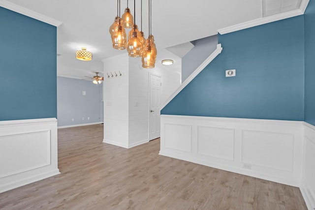 unfurnished dining area featuring a ceiling fan, a wainscoted wall, light wood finished floors, ornamental molding, and a decorative wall