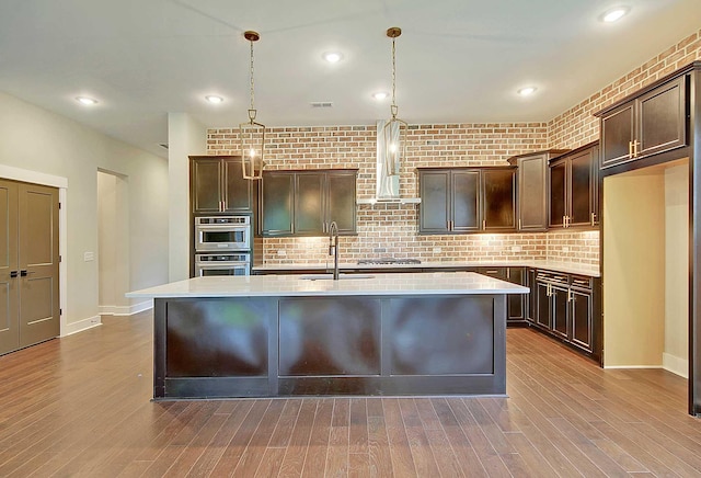 kitchen with wood-type flooring, pendant lighting, a center island with sink, and dark brown cabinets