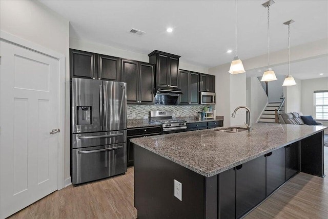 kitchen featuring appliances with stainless steel finishes, a kitchen island with sink, sink, decorative light fixtures, and dark stone countertops