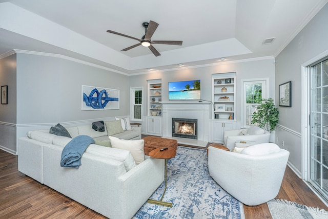 living room featuring dark wood-type flooring, built in shelves, ceiling fan, ornamental molding, and a tray ceiling