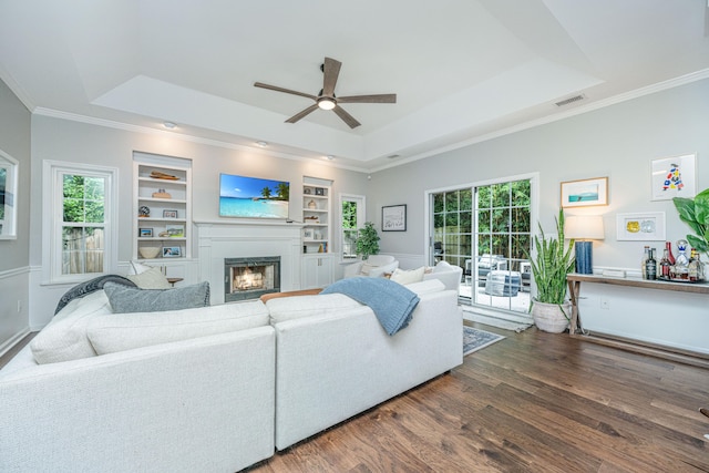 living room featuring built in shelves, ceiling fan, dark wood-type flooring, a raised ceiling, and ornamental molding