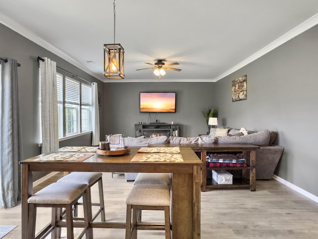 dining room featuring ornamental molding, light wood finished floors, a ceiling fan, baseboards, and a warm lit fireplace
