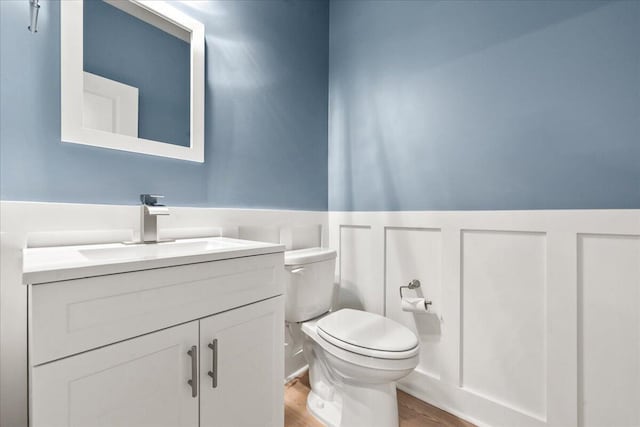 half bathroom with toilet, a decorative wall, wood finished floors, vanity, and wainscoting