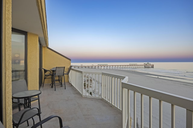 balcony at dusk featuring a water view and a beach view