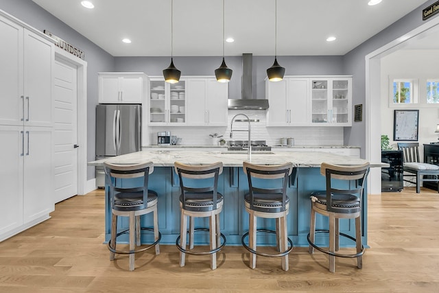 kitchen featuring a kitchen island with sink, light hardwood / wood-style flooring, hanging light fixtures, and wall chimney range hood