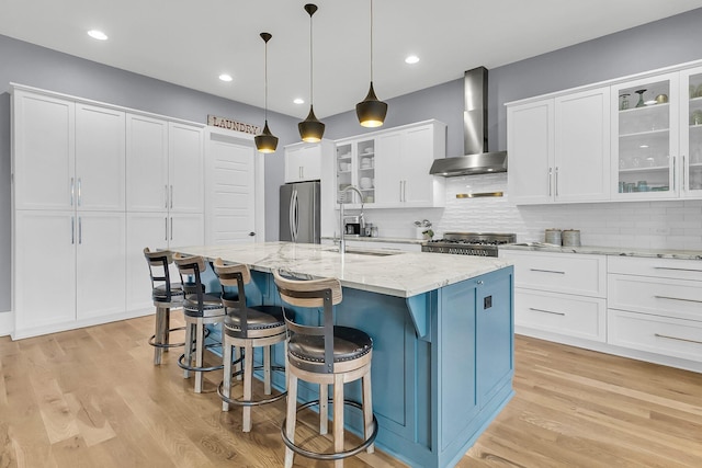 kitchen with appliances with stainless steel finishes, wall chimney exhaust hood, white cabinets, hanging light fixtures, and an island with sink