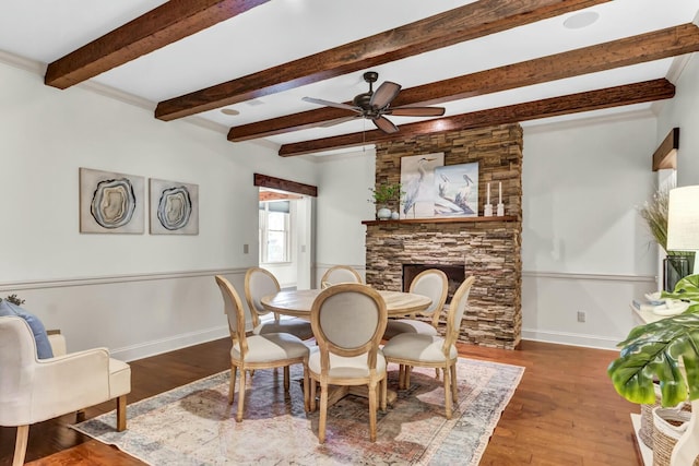 dining space featuring beam ceiling, wood finished floors, a stone fireplace, baseboards, and ceiling fan