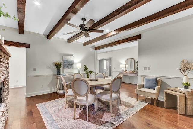 dining space with wood finished floors, baseboards, ceiling fan, a stone fireplace, and beamed ceiling