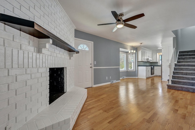 unfurnished living room featuring light wood finished floors, baseboards, stairway, a brick fireplace, and ceiling fan with notable chandelier