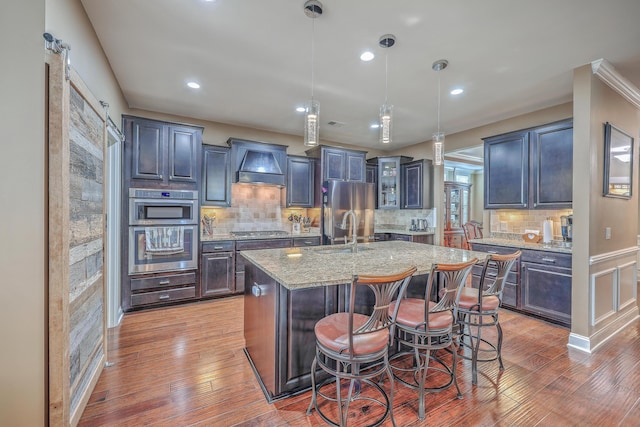 kitchen featuring stainless steel appliances, light stone counters, custom exhaust hood, a center island with sink, and pendant lighting