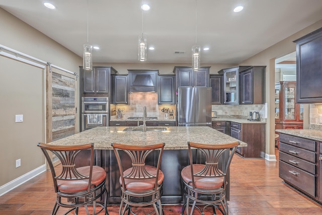 kitchen with stainless steel appliances, premium range hood, dark brown cabinets, a barn door, and a center island with sink