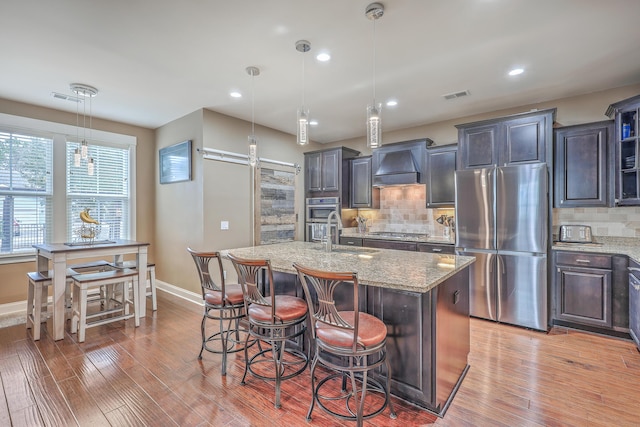 kitchen featuring a center island with sink, stainless steel appliances, hanging light fixtures, custom range hood, and sink