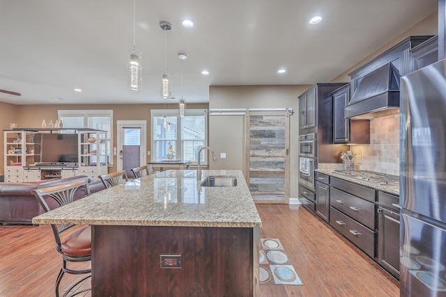 kitchen featuring light stone countertops, custom exhaust hood, stainless steel appliances, a kitchen island with sink, and sink